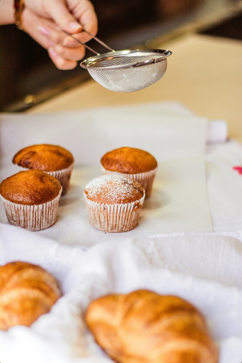 Baker dusting powdered sugar over muffins and croissants